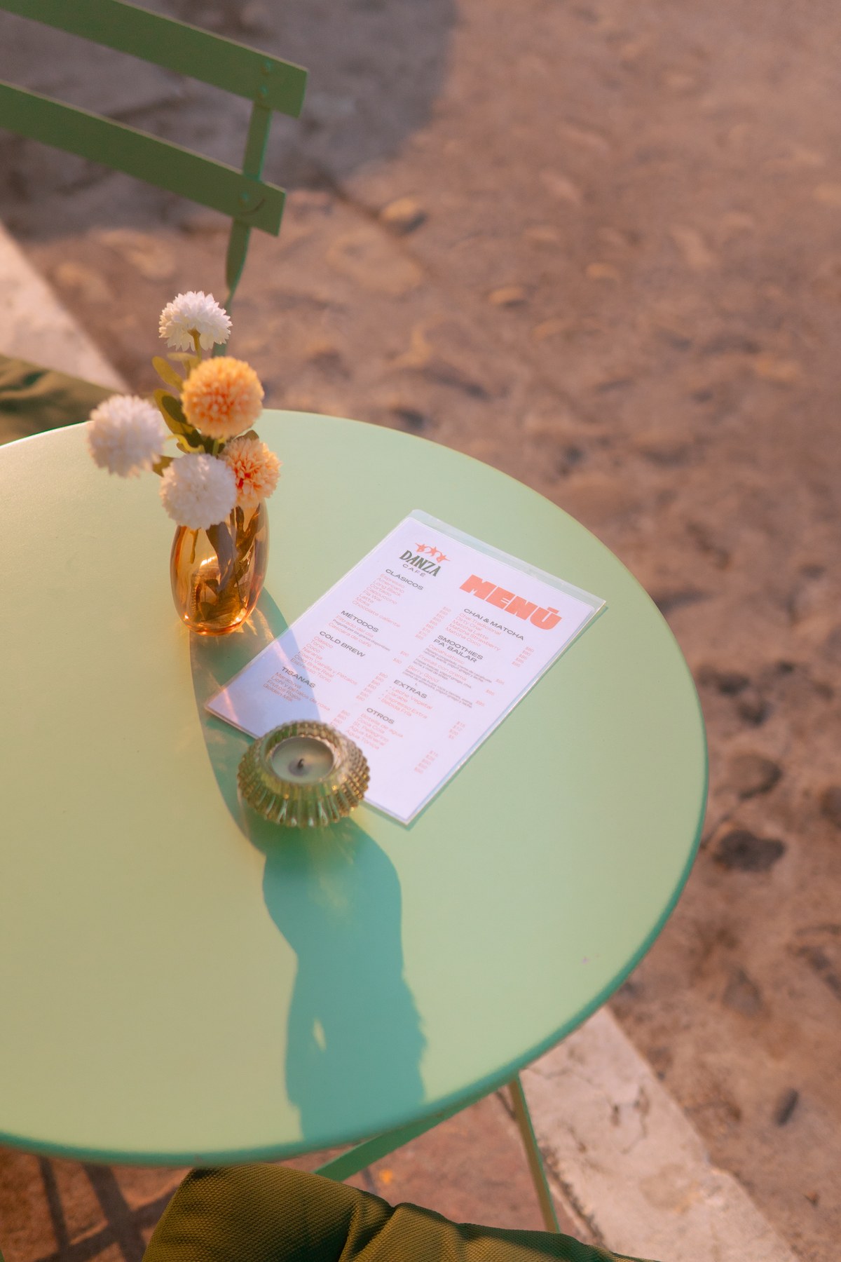 Top-down flatlay of green round bistro table in golden hour light with printed Danza menu, amber flower vase, and glass candleholder